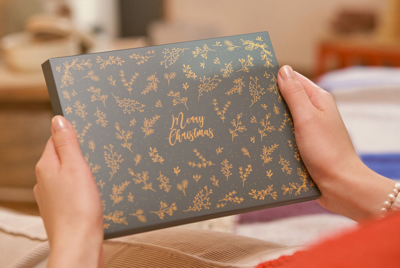Hände halten eine dunkelgraues Geschenk mit goldfarbenem, weihnachtlichem Muster und der Aufschrift "Merry Christmas"