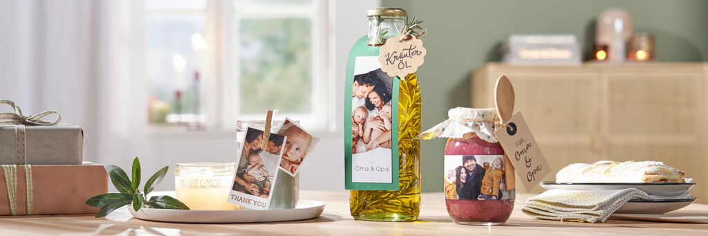 Verschiedene liebevoll gestaltete Geschenke und persönliche Erinnerungen auf einem Tisch: eine Flasche Öl, ein Glas Marmelade, ein Fotobuch und eine brennende Kerze. Alles präsentiert in warmem, einladendem Licht