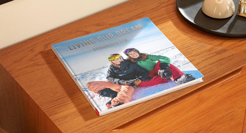 Zwei Personen mit Skiausrüstung sitzen auf einem Schneehügel und lächeln. Auf dem Tisch liegt ein CEWE Fotobuch.