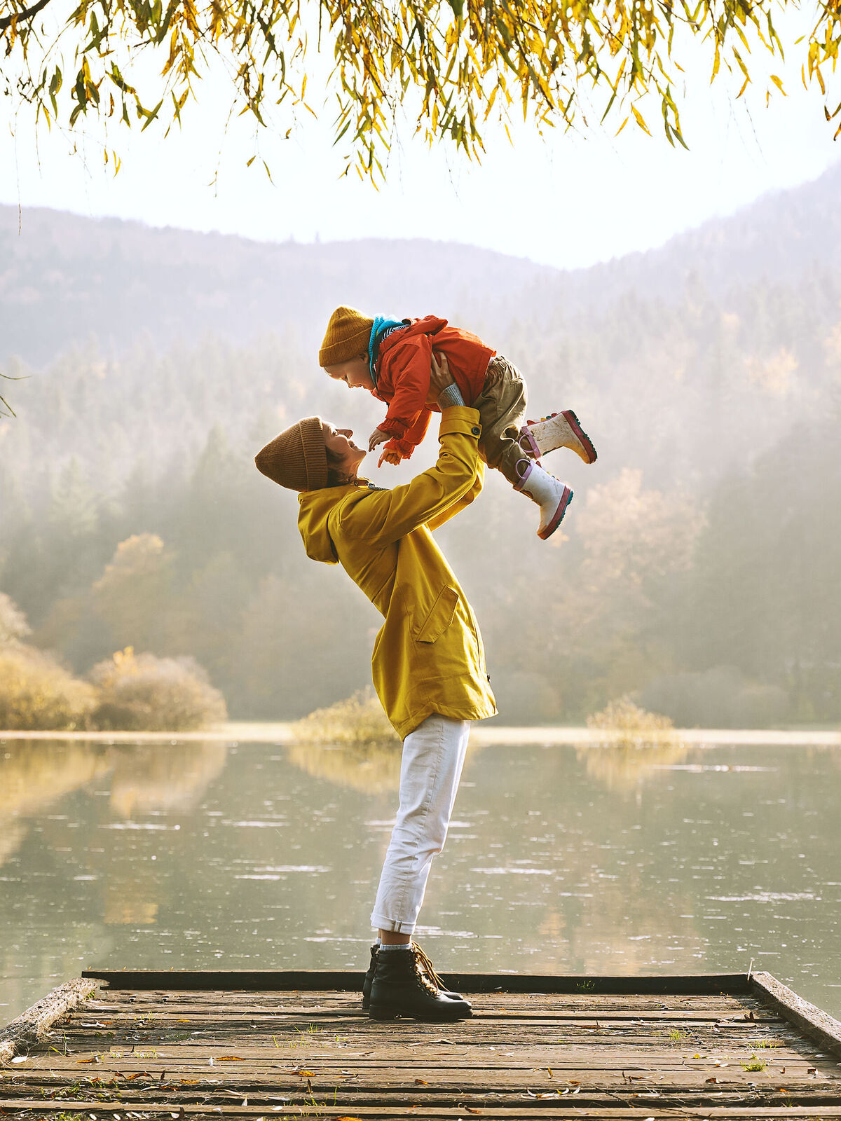 Ein fröhlicher Erwachsener hebt ein Kind in die Luft, vor einer malerischen Herbstlandschaft mit Wasser im Hintergrund, harmonische Farben.